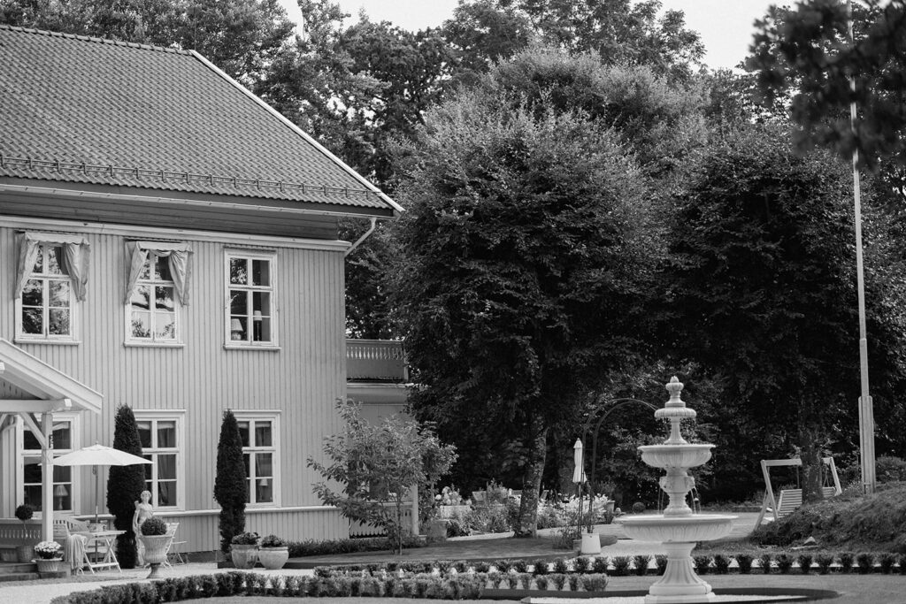 Black and white image of a house with a garden and a small fountain in front